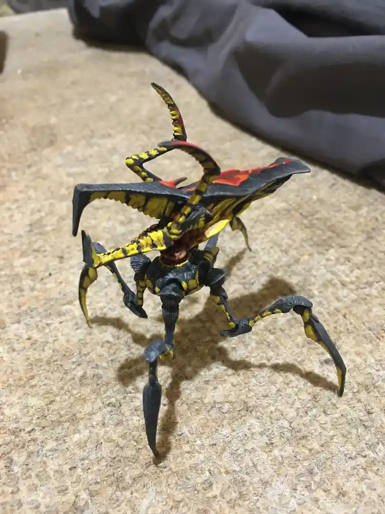 A hand-painted scorpion toy figure, predominantly black with yellow and red accents, is posed on a beige carpet. The…