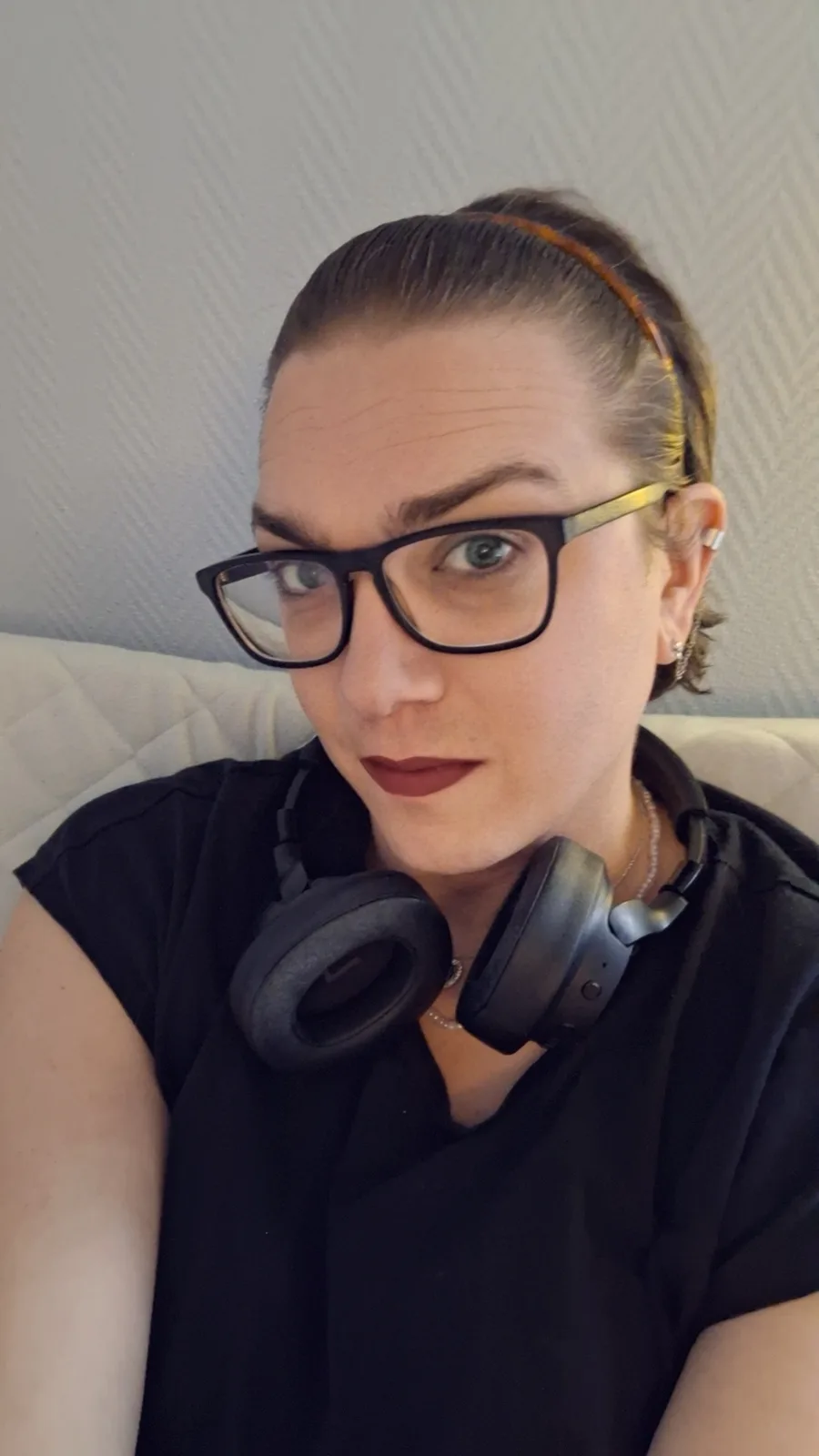 A person with glasses and headphones around their neck stares directly at the camera with a neutral expression, seated on a…