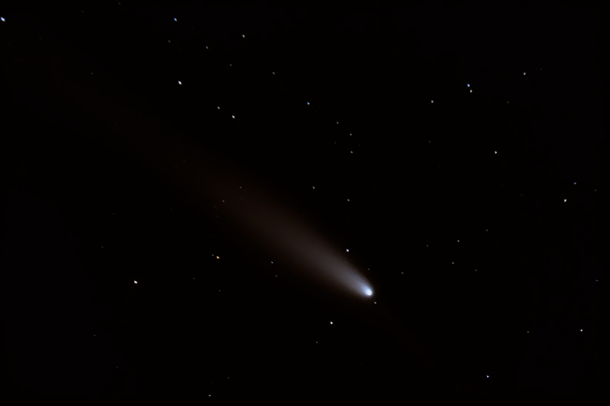 A celestial image shows a comet with a bright nucleus and a long, diffuse tail streaking across a dark sky dotted with…