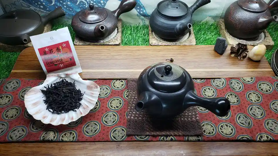 A wooden table displays a black ceramic teapot on a woven mat, next to a white seashell holding dark loose-leaf tea. Behind…