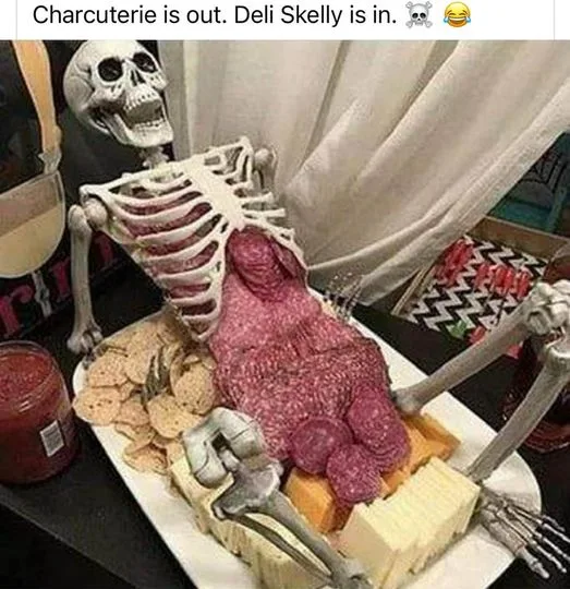 A plastic skeleton is posed reclining on a platter filled with deli meats, cheese, and crackers, with its ribcage open to…