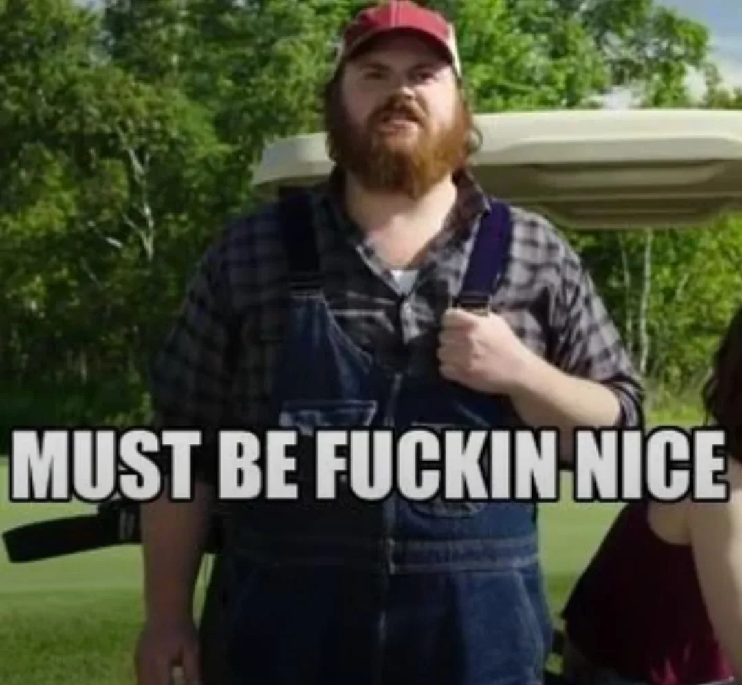 A man with a red cap and beard, wearing overalls and a plaid shirt, stands on a golf course holding a golf bag, looking…