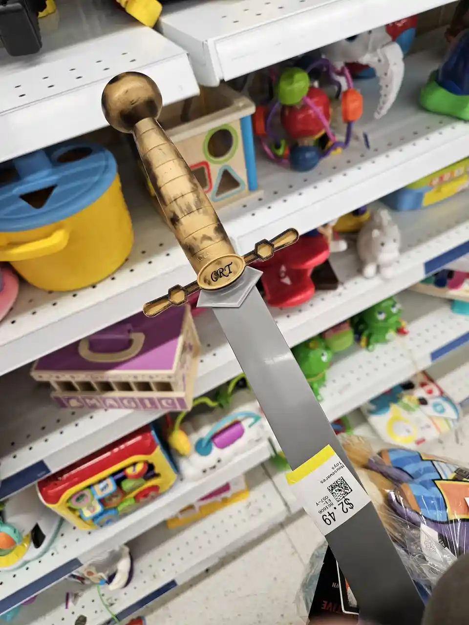 A close-up photo of a toy sword with a gold-colored hilt and a silver blade, displayed on a store shelf among other…