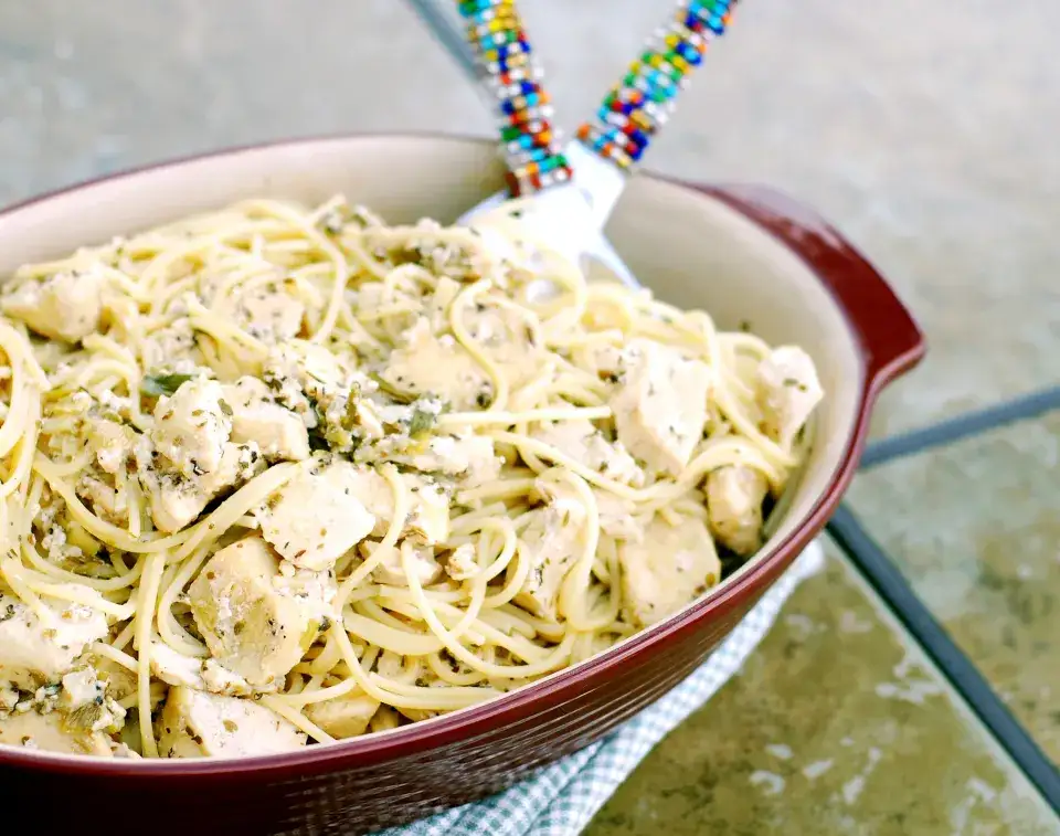 A close-up photo of a red ceramic baking dish filled with creamy chicken pasta, garnished with herbs and cheese, resting on…