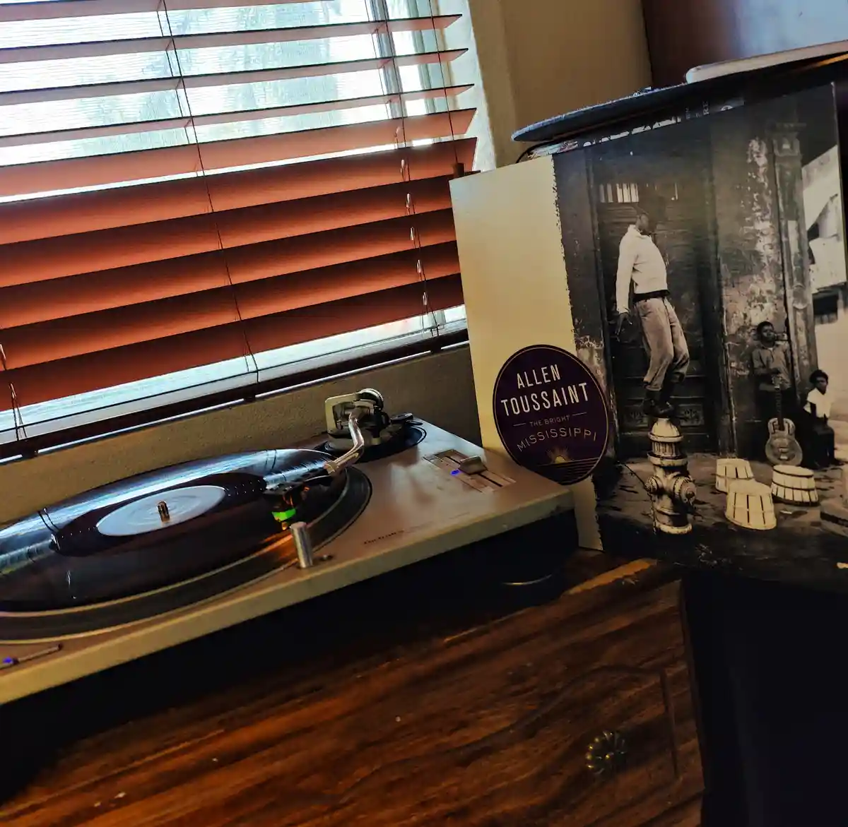 A vinyl record player sits on a wooden surface near a window with horizontal blinds, next to the album cover for 'The Bright…
