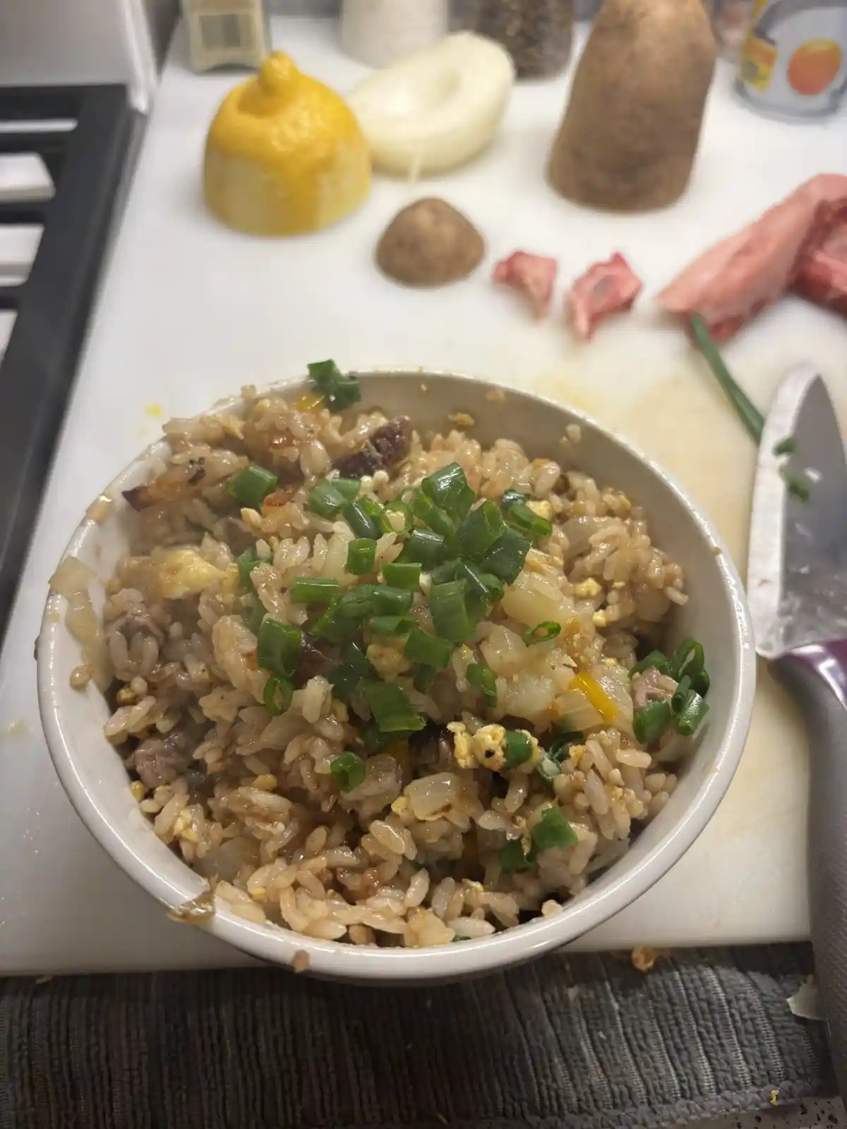 A close-up photo shows a bowl of fried rice topped with chopped green onions, sitting on a white cutting board. In the…