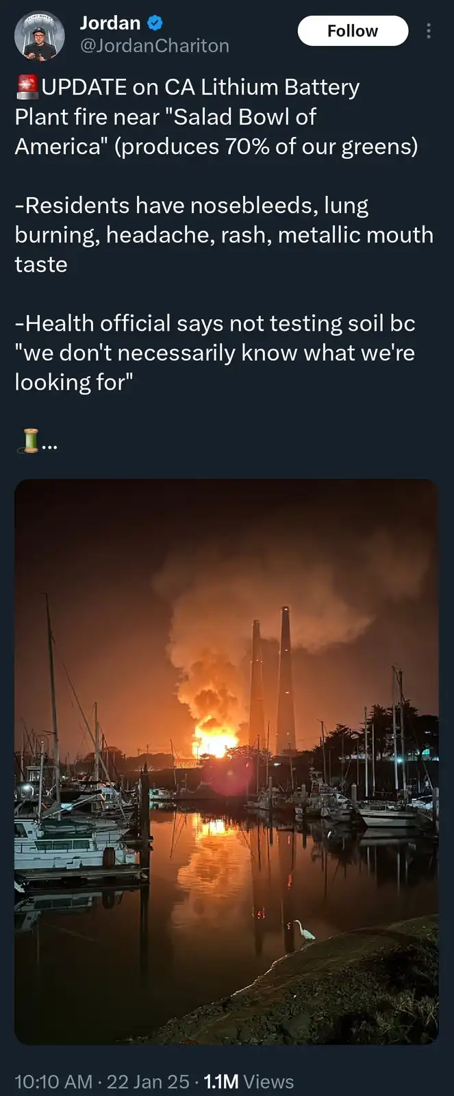 A nighttime photo shows a large fire and smoke plume erupting from an industrial facility with tall smokestacks, viewed…