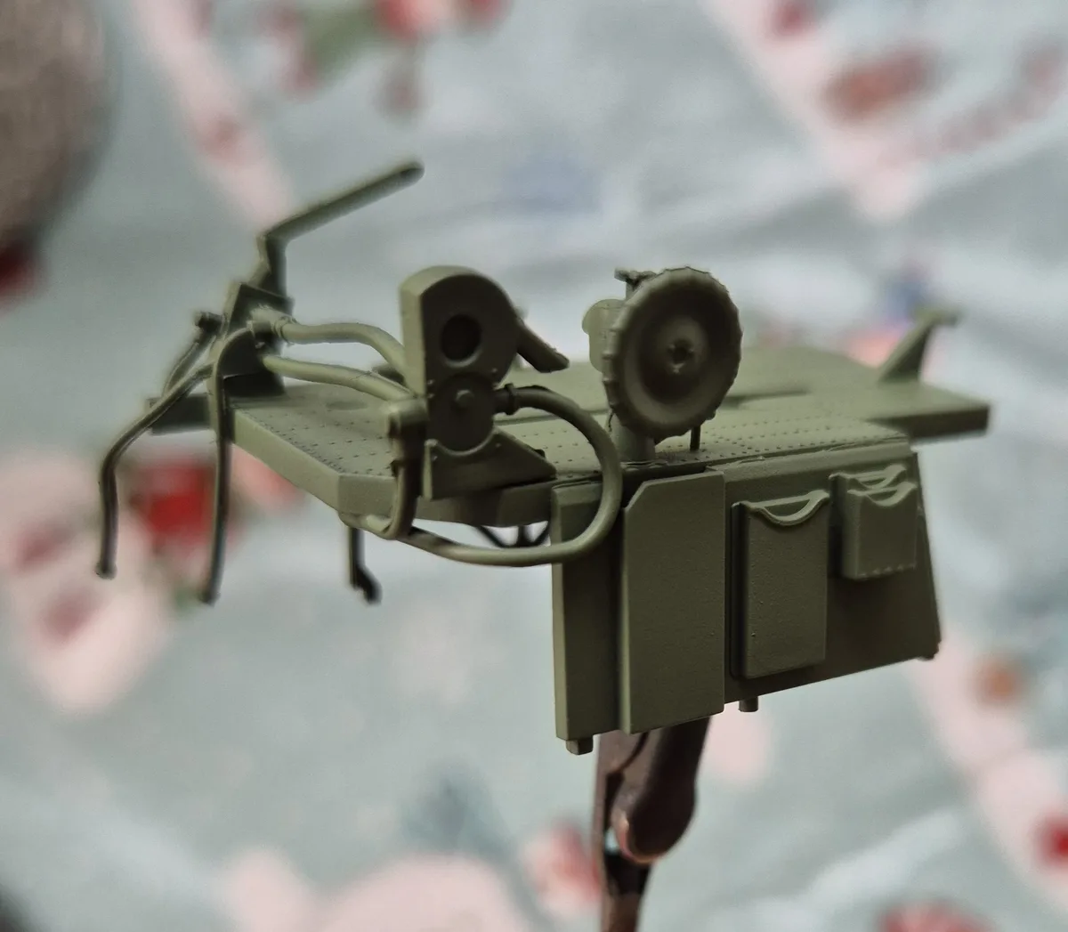 A close-up photo shows a miniature olive-drab military vehicle model, possibly a tank or armored car, mounted on a clamp…