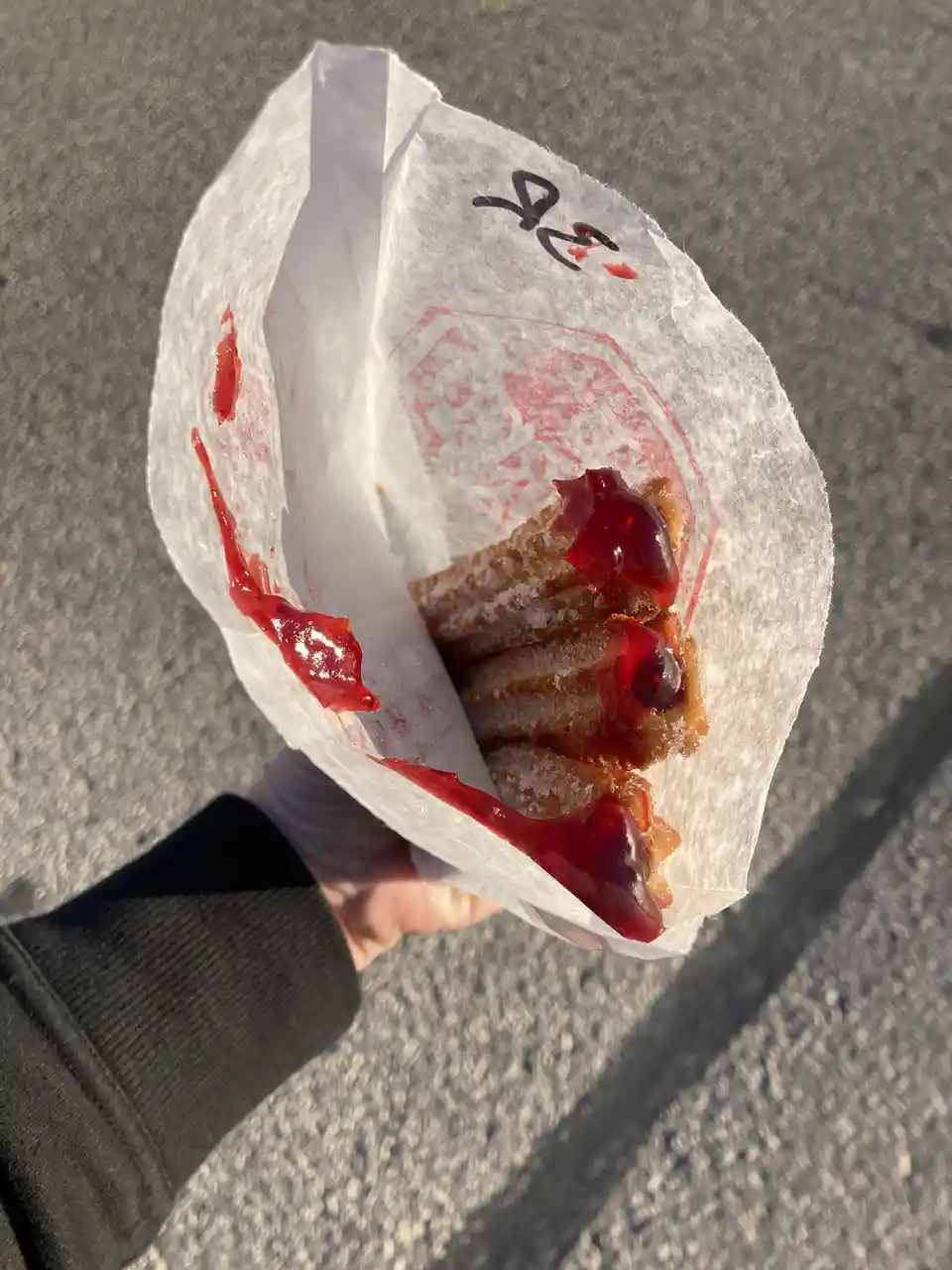 A hand holds a white paper wrapper containing sugared churros drenched in a thick red sauce that drips down the sides…