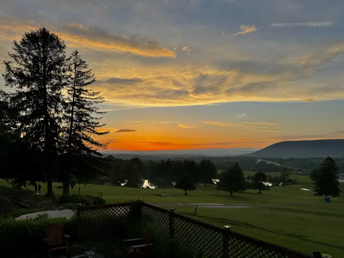 A scenic photograph of a sunset over a golf course with silhouetted trees and a wooden lattice fence in the foreground. Two…
