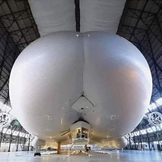 a low-angle shot of the rear section of a large silver airship inside a hangar, with its rounded, bulbous shape and visible…