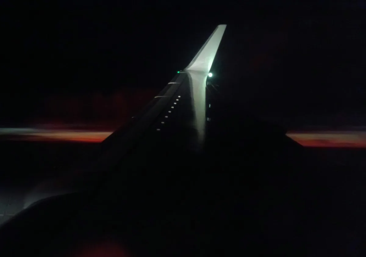 A blurry photo taken from an airplane window at night, showing the illuminated wing stretching into the dark sky with…