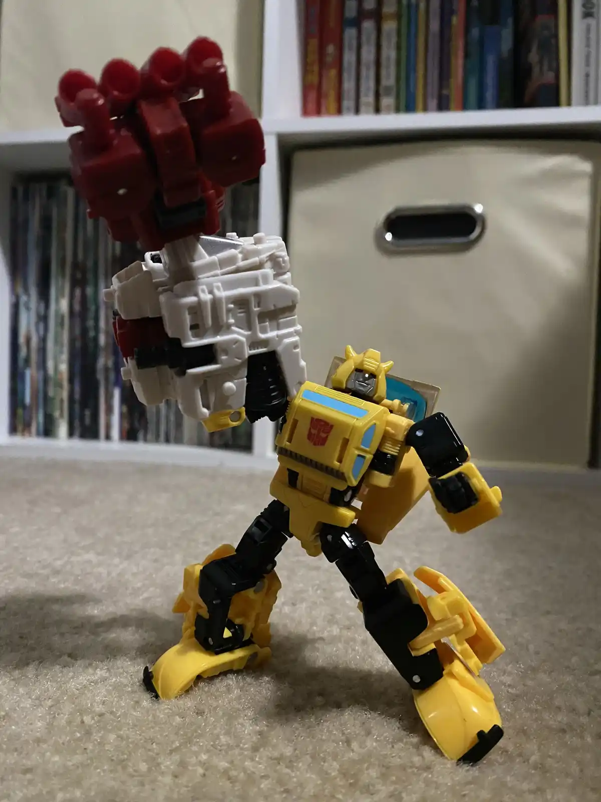 A yellow and black Bumblebee Transformers toy stands on a beige carpet, holding up a large red and white robot head that…