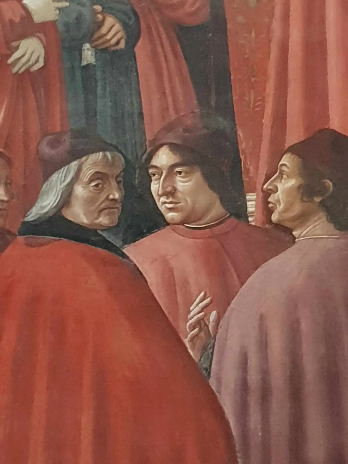 A close-up of a Renaissance painting shows three men in red and dark robes, one with gray hair looking away, another in the…
