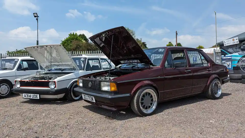 Two classic Volkswagen cars with their hoods open are parked side-by-side on gravel under a blue sky. The maroon car in the…