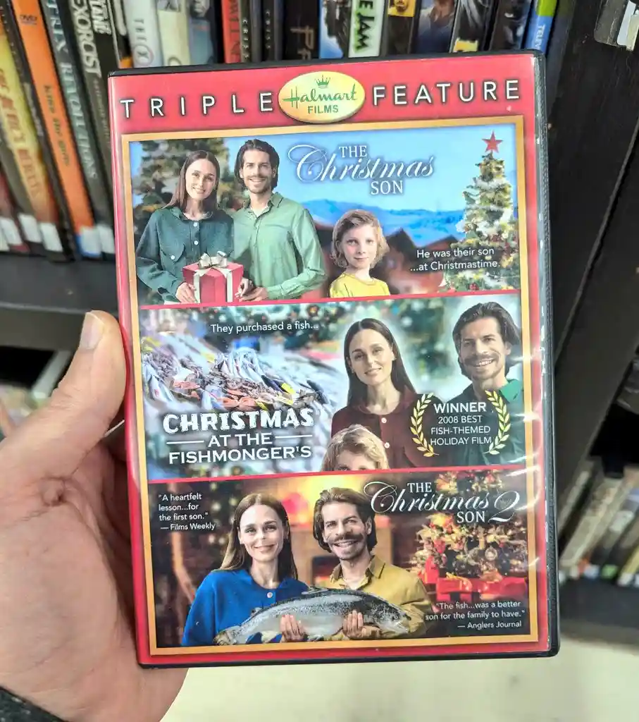 A hand holds a DVD case labeled 'Triple Feature' from 'HalMart Films,' which humorously presents three Christmas-themed…