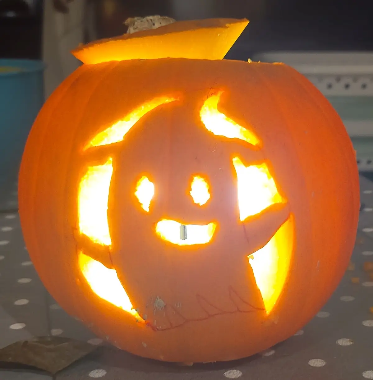 A carved Halloween pumpkin with a glowing face, featuring horns, eyes, and a wide grin with a single visible tooth. The…