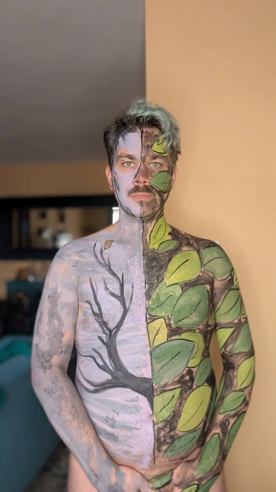 A person stands shirtless with their body painted to depict a split tree: one half is gray with bare branches, the other…
