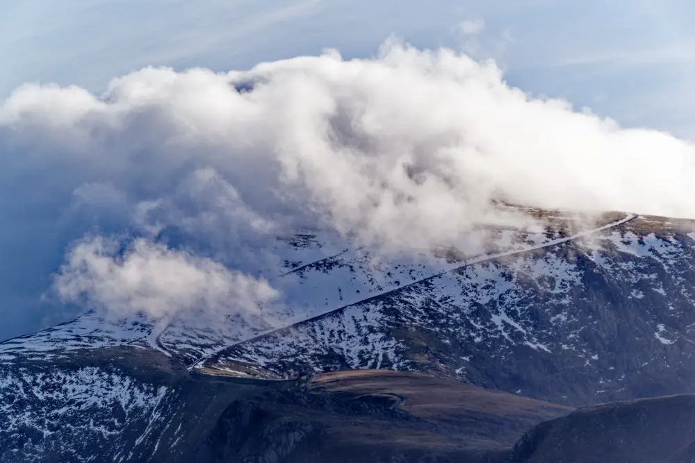 A scenic mountain landscape with snow-dusted peaks and a winding road partially obscured by thick white clouds drifting…