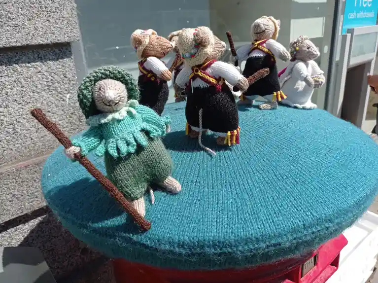 Several hand-knitted mouse dolls dressed in traditional folk costumes are arranged as if dancing on a large, teal knitted…