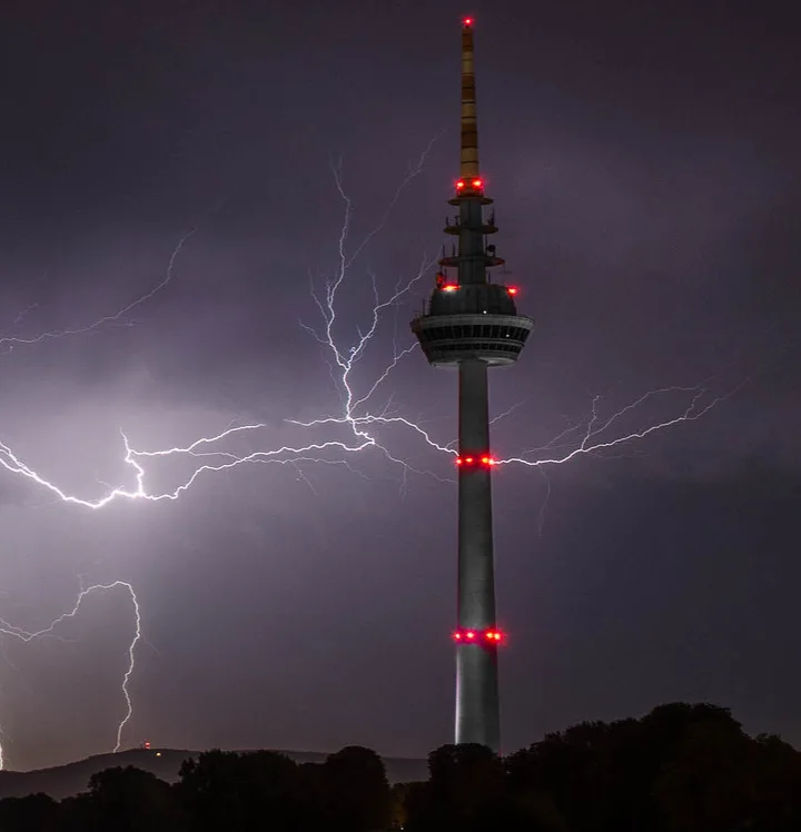 A tall, slender tower with red lights at intervals stands against a dark, stormy sky filled with bright lightning bolts. The…