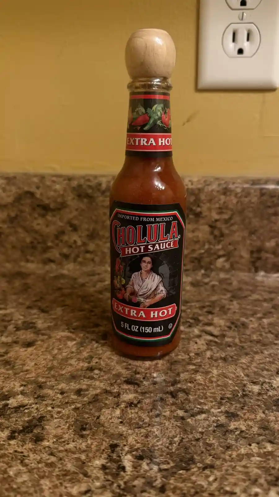 A bottle of Cholula Extra Hot hot sauce sits on a speckled brown countertop, with a yellow wall and white electrical outlet…
