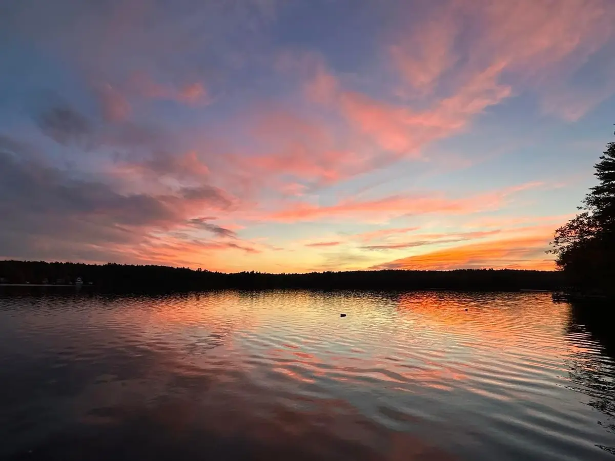 A serene sunset over a calm lake, with vibrant pink, orange, and purple clouds reflected in the water. The horizon shows a…
