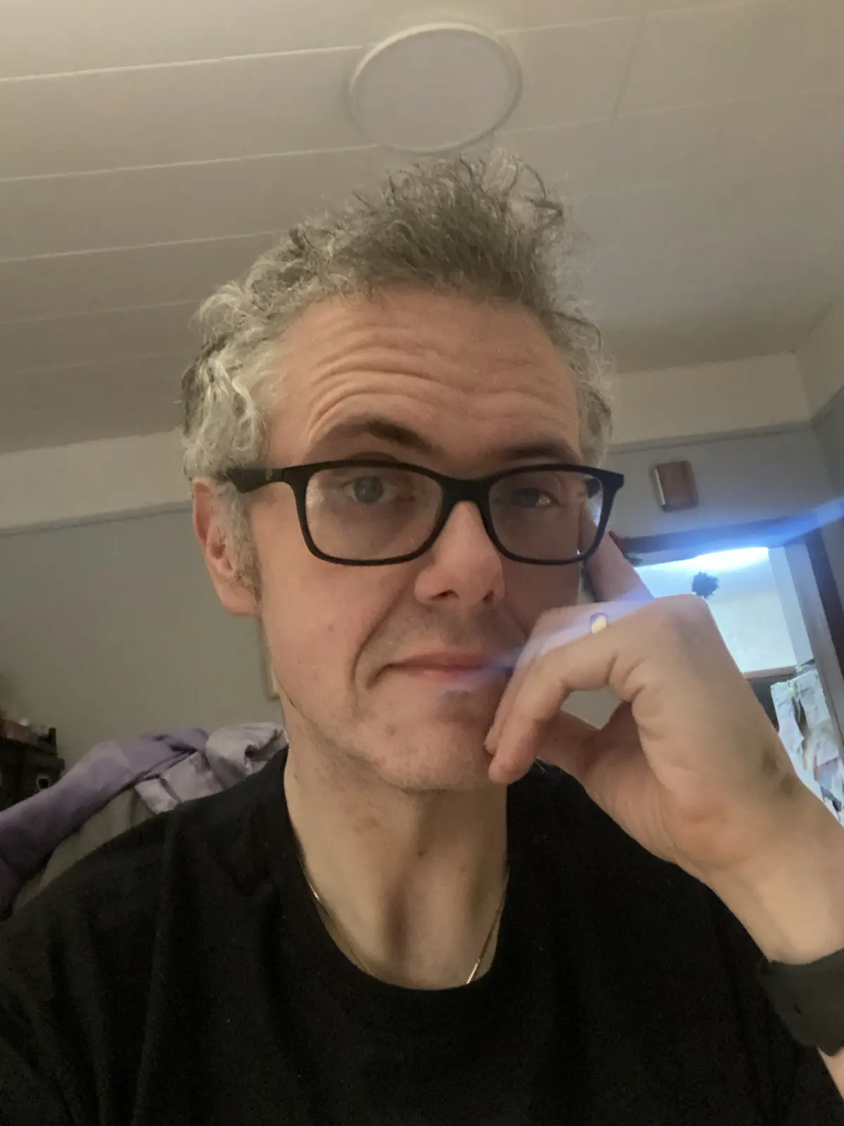 A man with curly gray hair and black-rimmed glasses looks directly at the camera with a smug expression while holding his…
