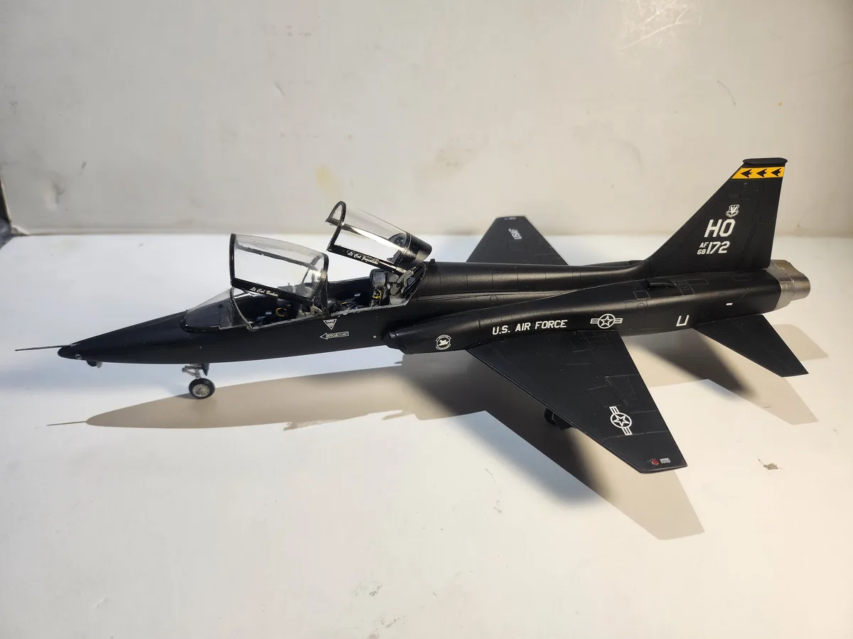 A black model of a U.S. Air Force jet, likely a T-38 Talon trainer, sits on a white surface with its canopy open. The…
