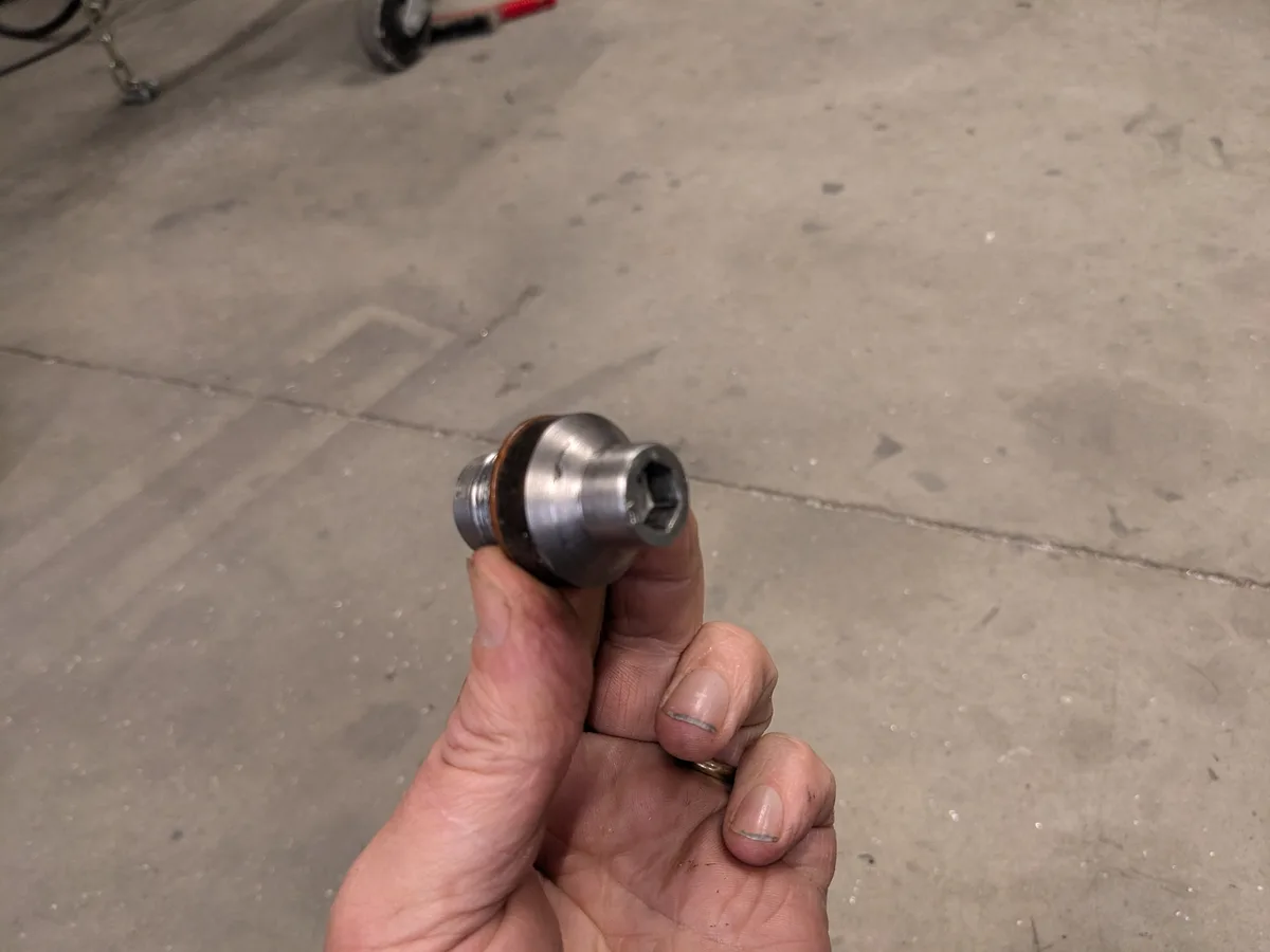 A hand holds a small, metallic bicycle repair tool with a hexagonal socket end, against a concrete floor background. The…