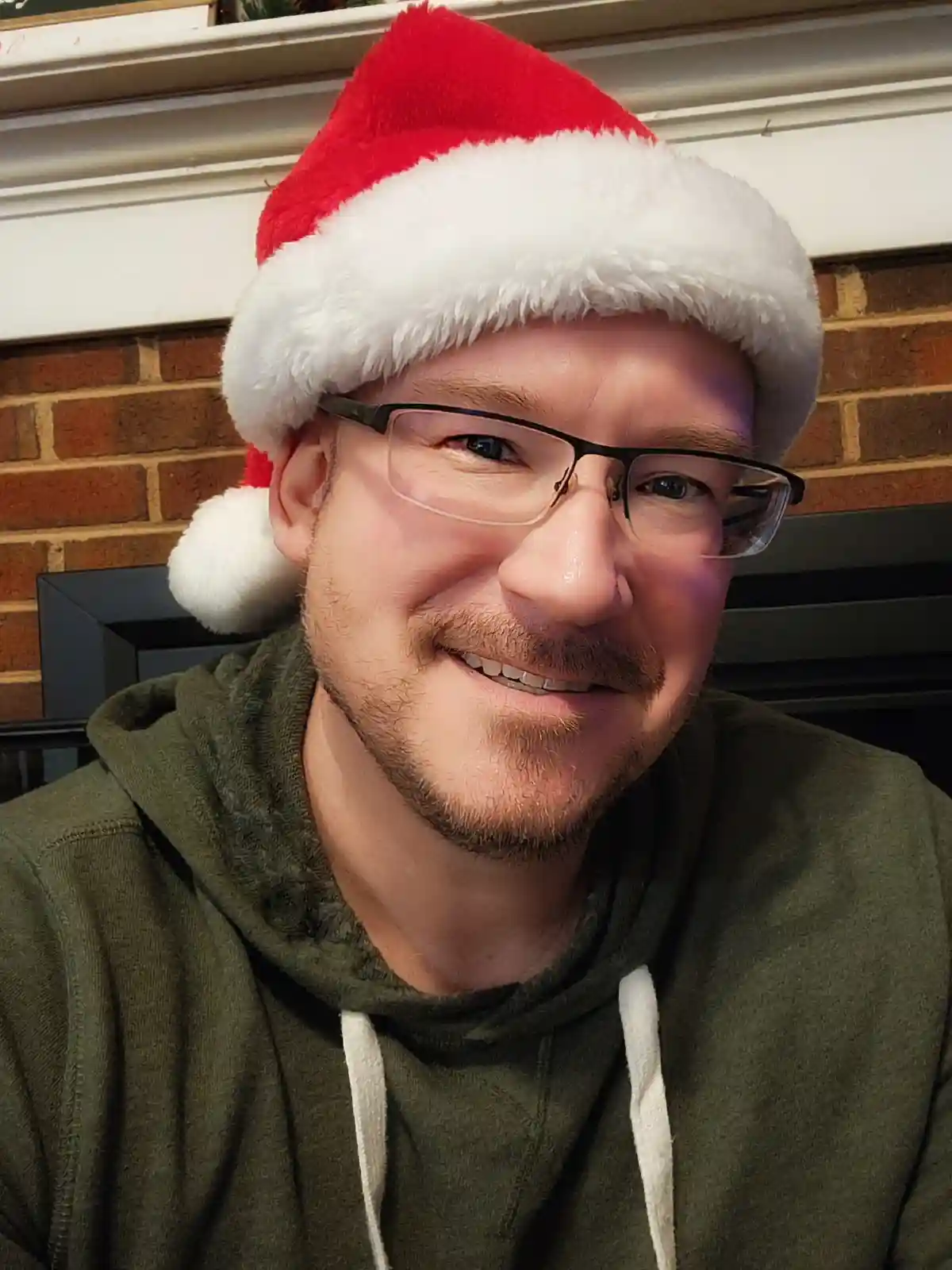 A man with glasses and a goatee smiles while wearing a red and white Santa hat and an olive green hoodie, seated in front of…