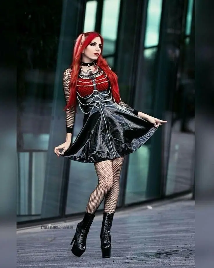 A woman with long red hair and gothic fashion, wearing a black latex skirt, fishnet stockings, and high-heeled boots, poses…