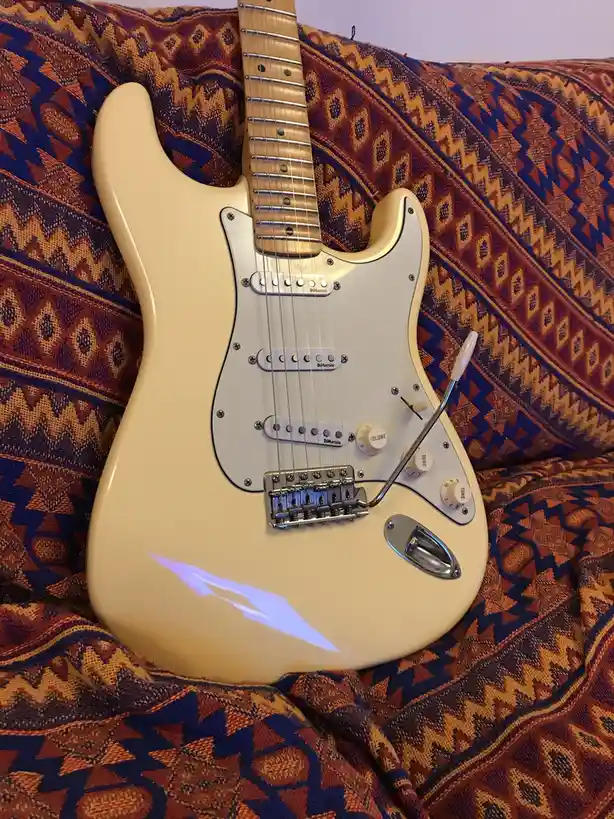 A cream-colored electric guitar, resembling a Fender Stratocaster with DiMarzio pickups and a white pickguard, rests on a…