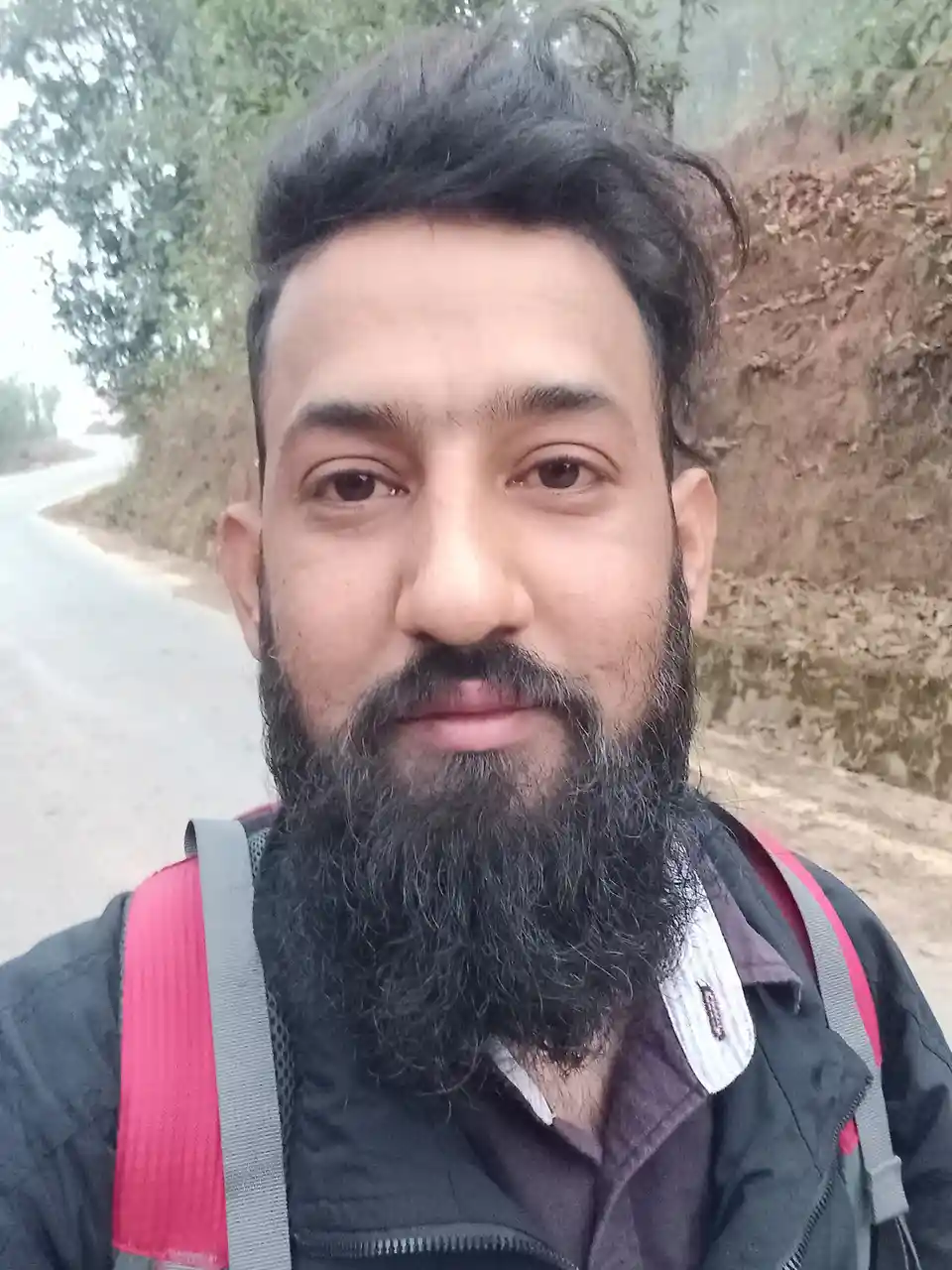 A man with a full beard and dark hair takes a selfie outdoors on a road, wearing a backpack with red and gray straps over a…