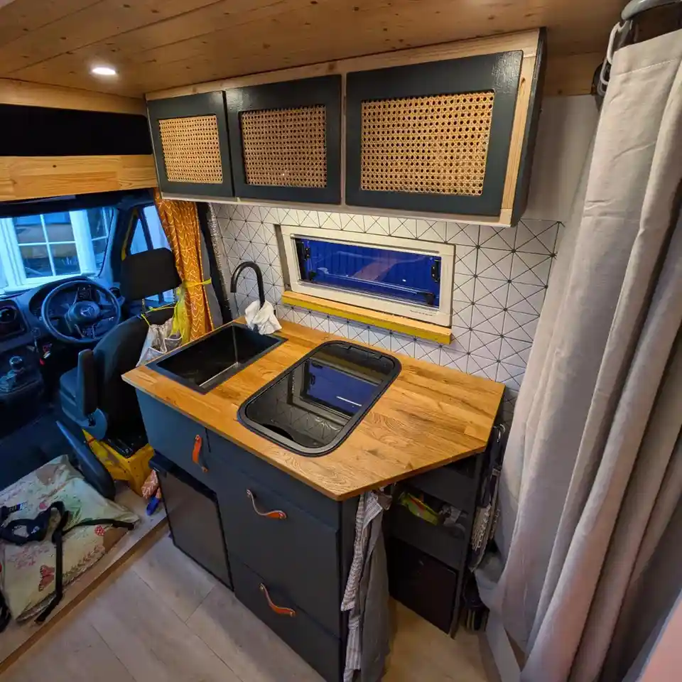 A cozy, custom-built kitchen inside a van features dark gray cabinets with woven cane fronts, a wooden countertop with two…