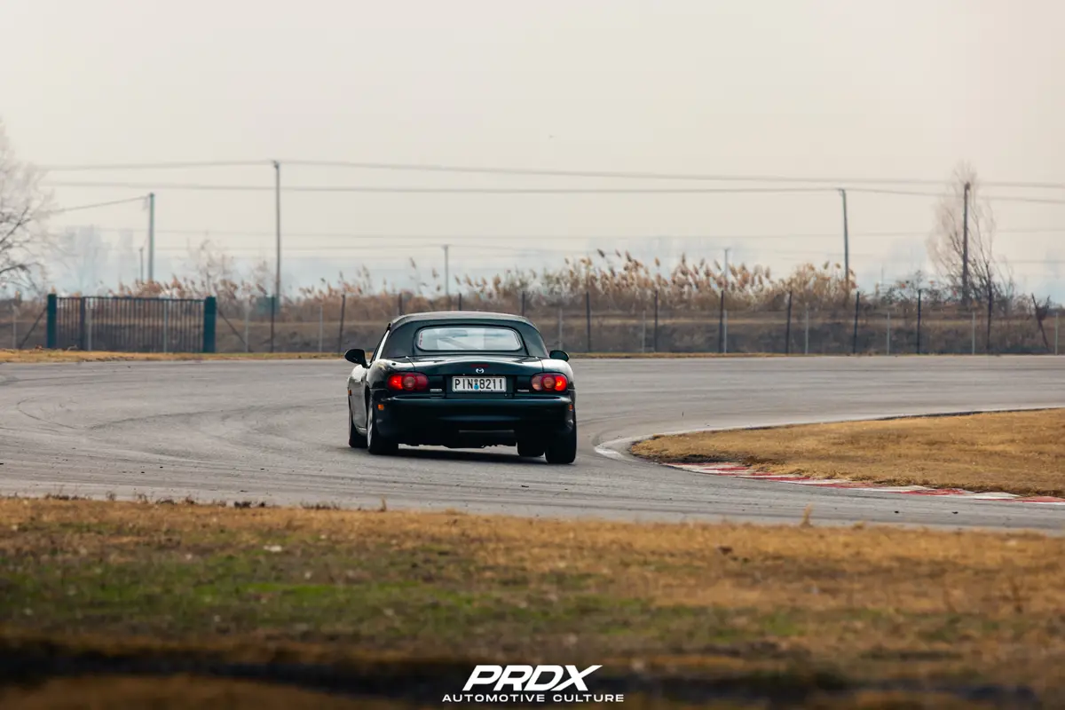 A black Mazda Miata convertible is shown from behind, drifting around a corner on an asphalt racetrack with red and white…