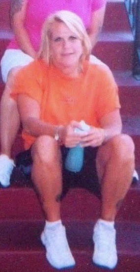 A woman with blonde hair sits on red stadium-style steps wearing an orange t-shirt, black shorts, and white socks. She holds…