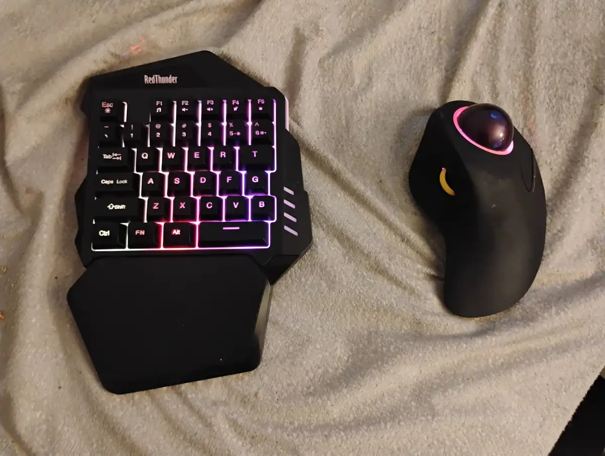 A compact, black RedThunder keyboard with purple backlighting and a built-in wrist rest sits next to a black trackball mouse…