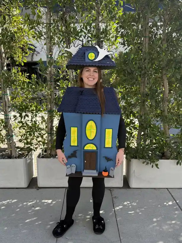 A smiling woman stands outdoors wearing a full-body Halloween costume shaped like a haunted blue house with a dark roof…