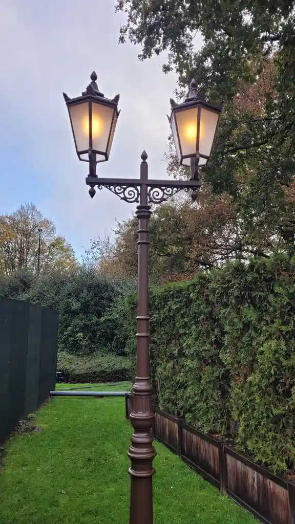 A vintage-style dual-lamp streetlight with ornate metalwork stands in a grassy path bordered by hedges and fences, its two…