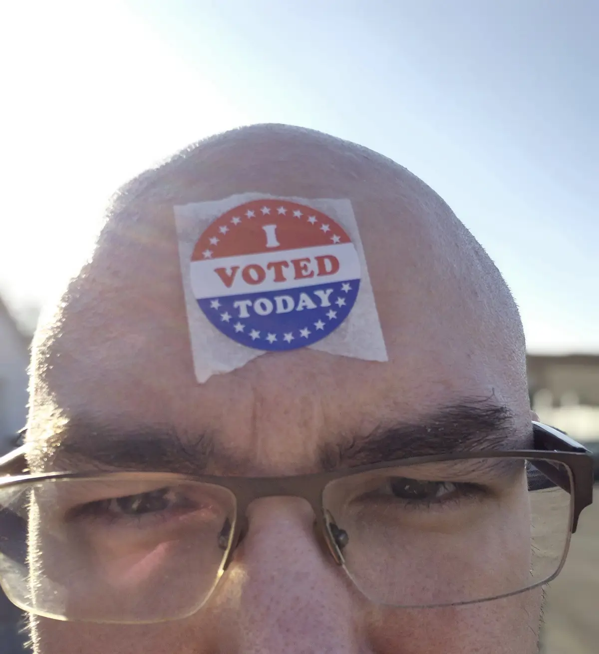 A close-up selfie of a man with glasses, showing a balding head with an 'I VOTED TODAY' sticker stuck directly to his…