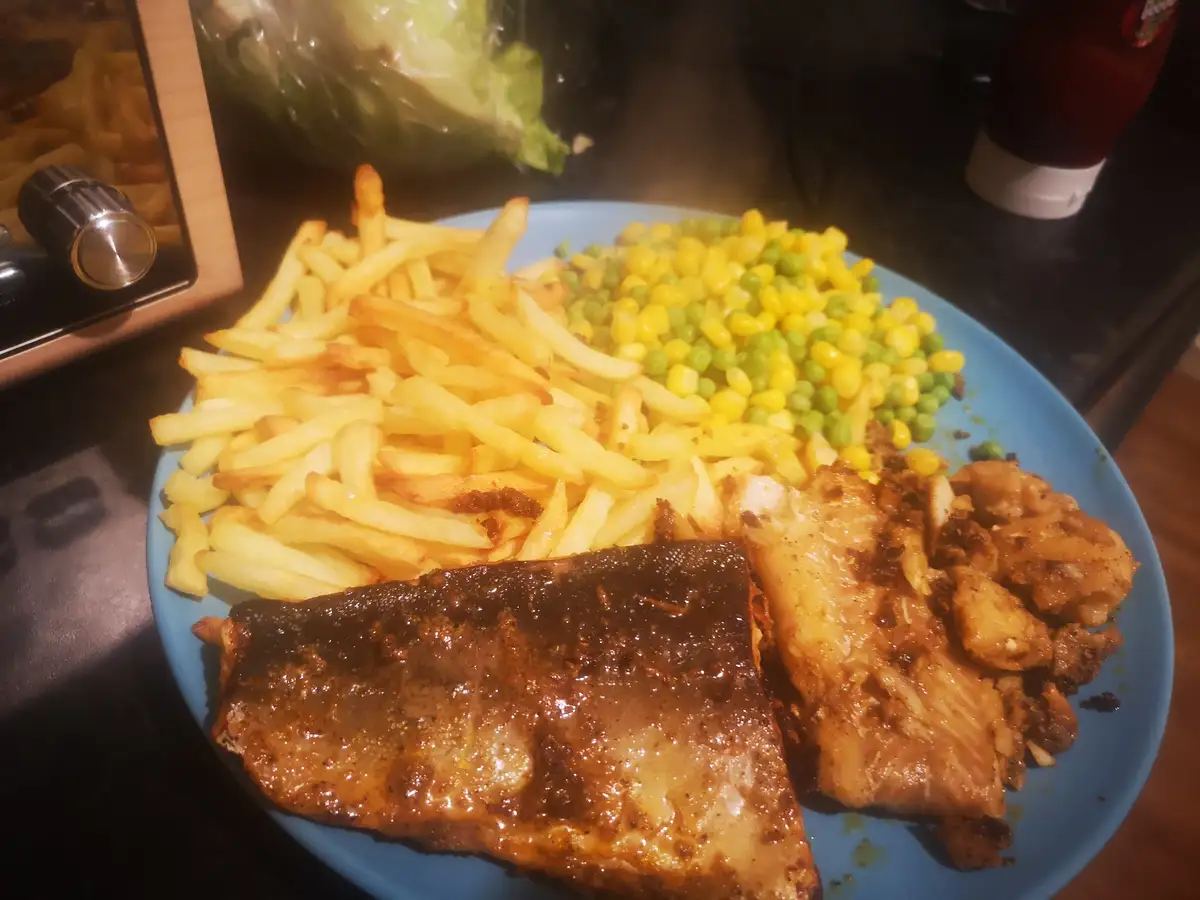 A blue plate holds a meal of golden-brown french fries, mixed corn and peas, and two pieces of seasoned, cooked fish. Steam…