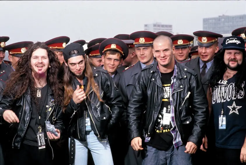 A group photo featuring several men in black leather jackets and casual attire, including one with long hair and another…