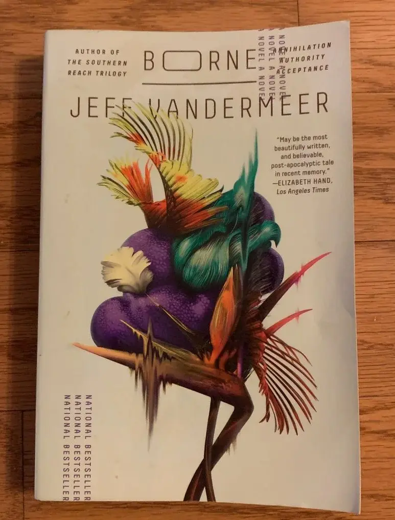 A book cover for Jeff VanderMeer's novel 'Borne' is photographed on a wooden surface. The cover features a surreal, colorful…