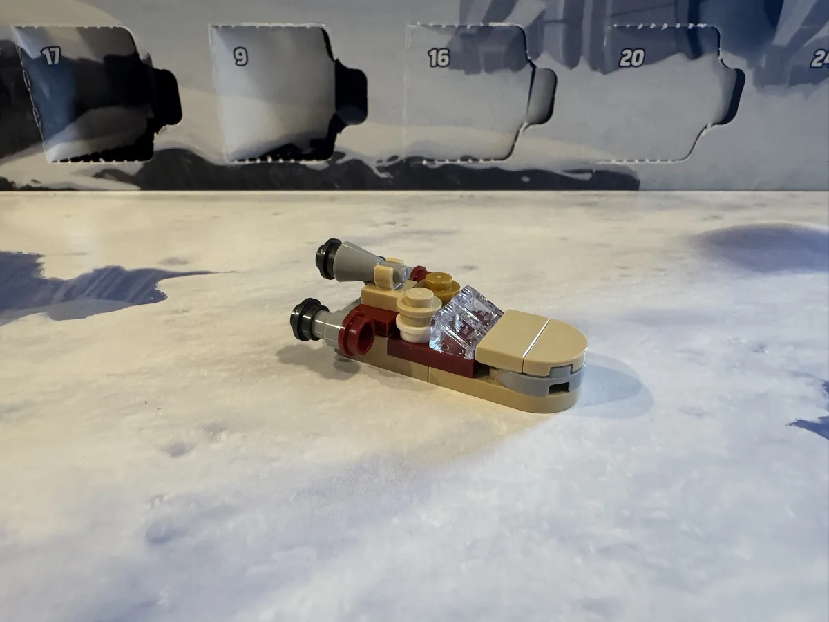 A small, beige, red, and gray LEGO spaceship model lies on a white, textured surface resembling snow or ice. Behind it, a…