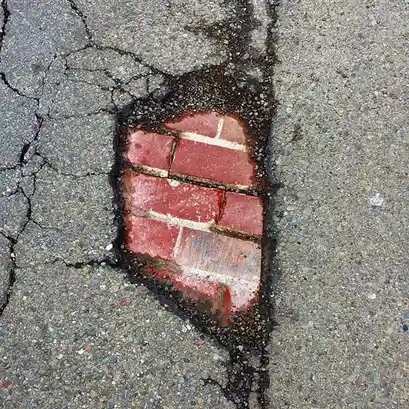 A close-up photo of a cracked asphalt road with a pothole shaped like a brick wall, revealing red bricks underneath. The…