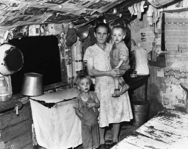 A black-and-white historical photo shows a woman holding a toddler while a young boy stands beside her in a severely…