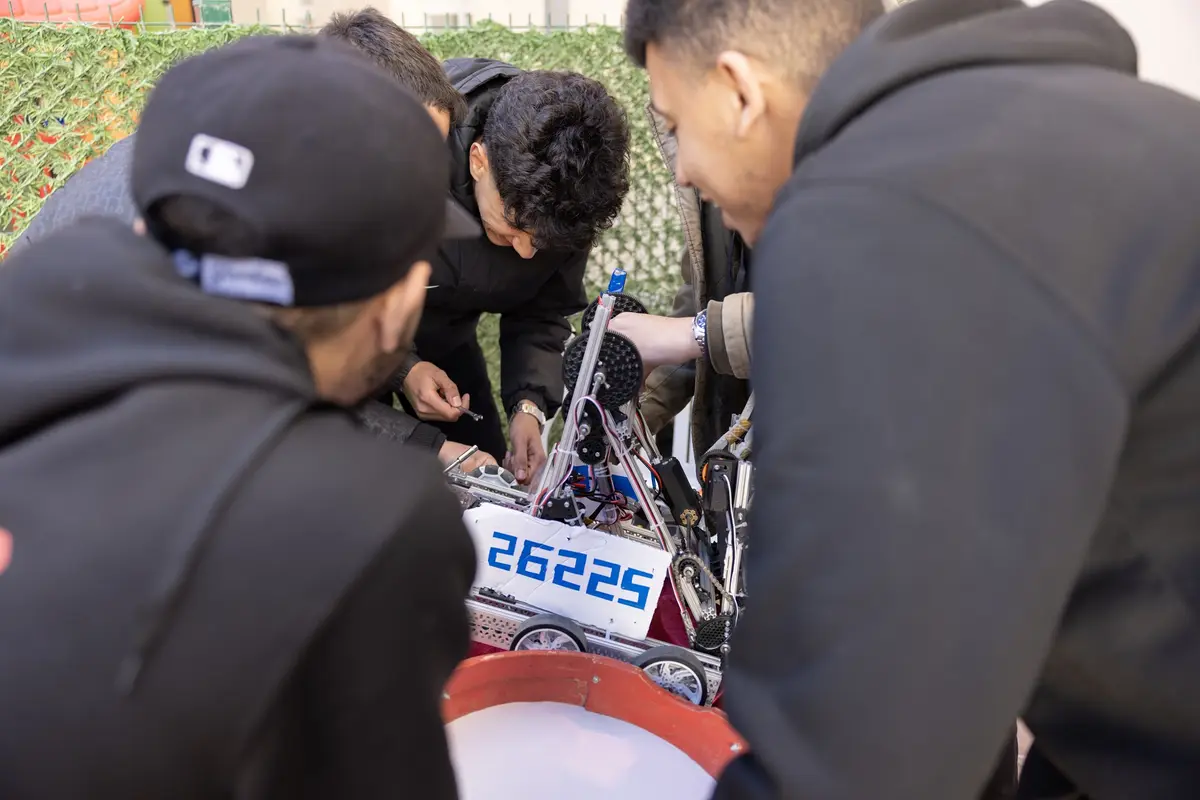 Four young men in hoodies and jackets are gathered around a small, wheeled robot with exposed gears and wires, intently…