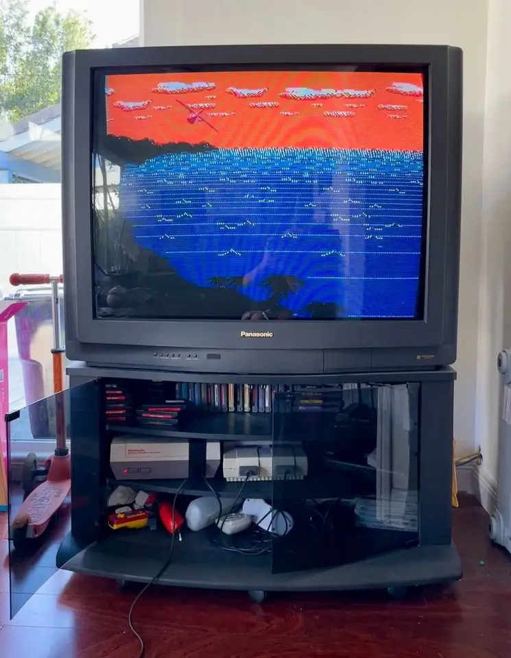 A large, old Panasonic CRT television displays a pixelated video game with a red sky and blue water, while the TV stand…