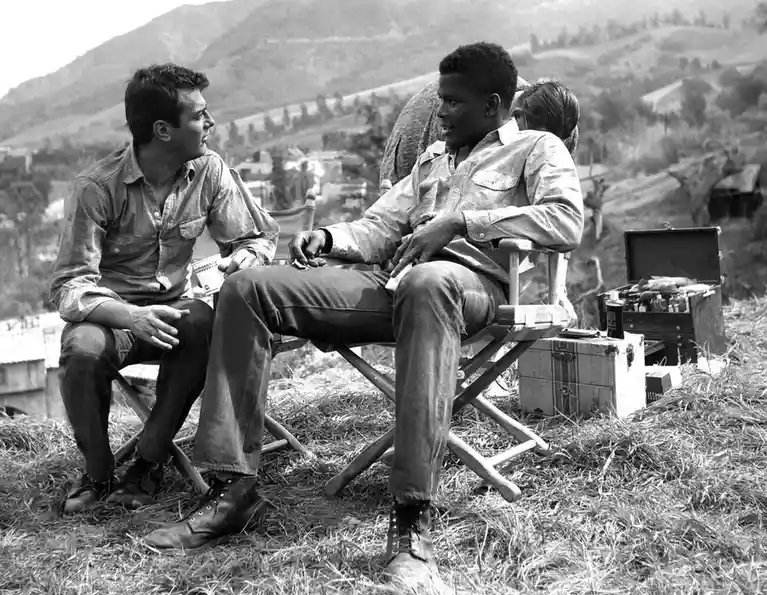 A black-and-white photo shows two men sitting in director's chairs outdoors on grassy terrain with hills in the background…